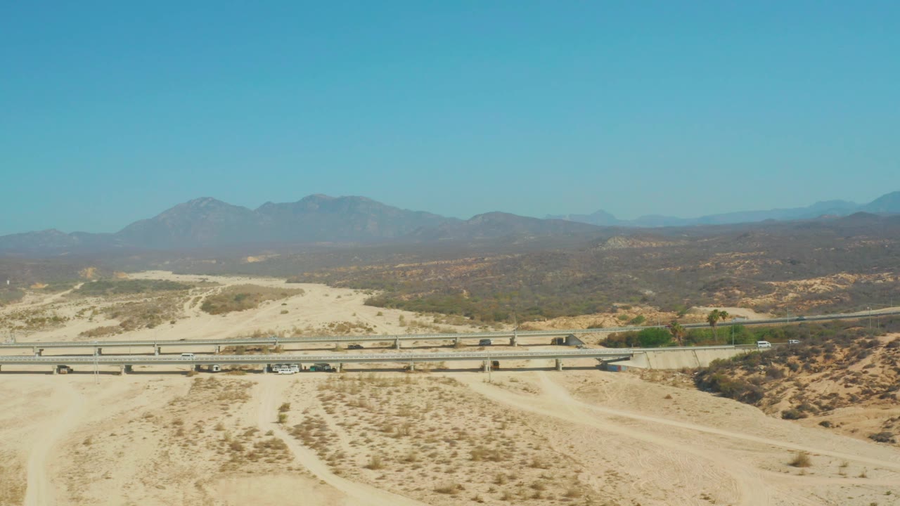 Aerial frame over El Tule Beach Los Cabos with the highway stretching across the lower scene, vehicles on the lanes, broad sandy ground, low vegetation, and no visible people.