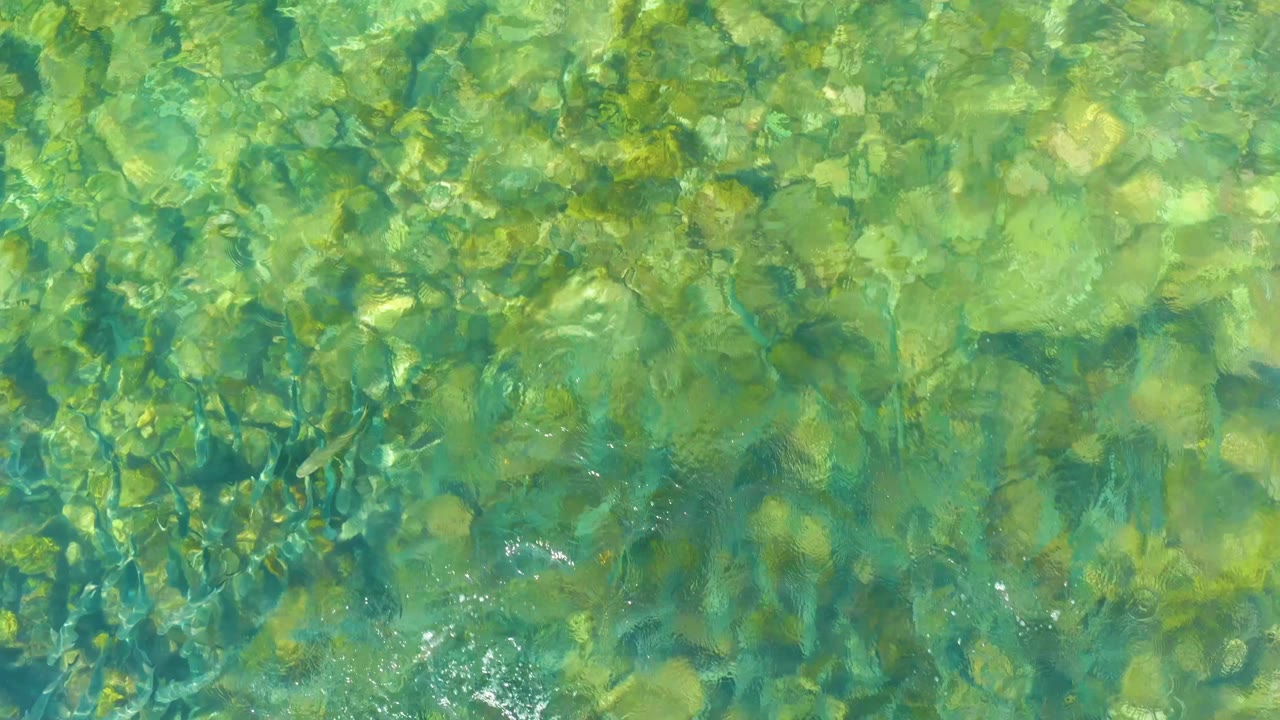 Green-blue water at El Tule Beach shows dense rock shapes below and a small bright patch of surface agitation near the lower center, with no visible people.
