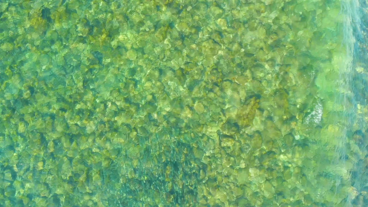 Clear turquoise water at El Tule Beach Los Cabos shows a dense bed of small rocks, with pale ripples crossing near the upper center and no visible people.