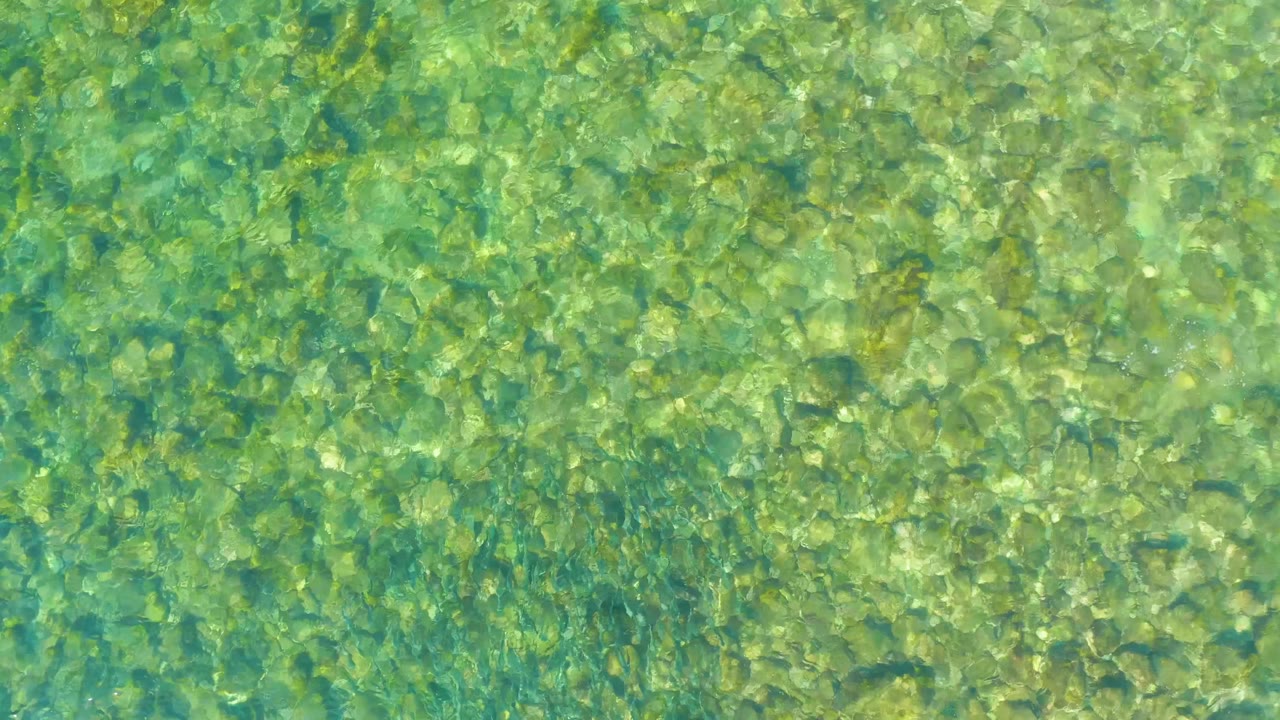 A high-angle frame of El Tule Beach Los Cabos water reveals yellow-green rocks, darker underwater patches, and a brighter ripple line with no visible people.