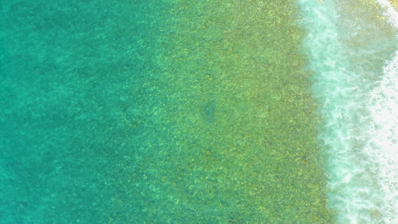 Aerial frame from El Tule Beach showing teal water on the left, pale green shallows through the center, and foamy surf pressed against the right edge, no visible people.