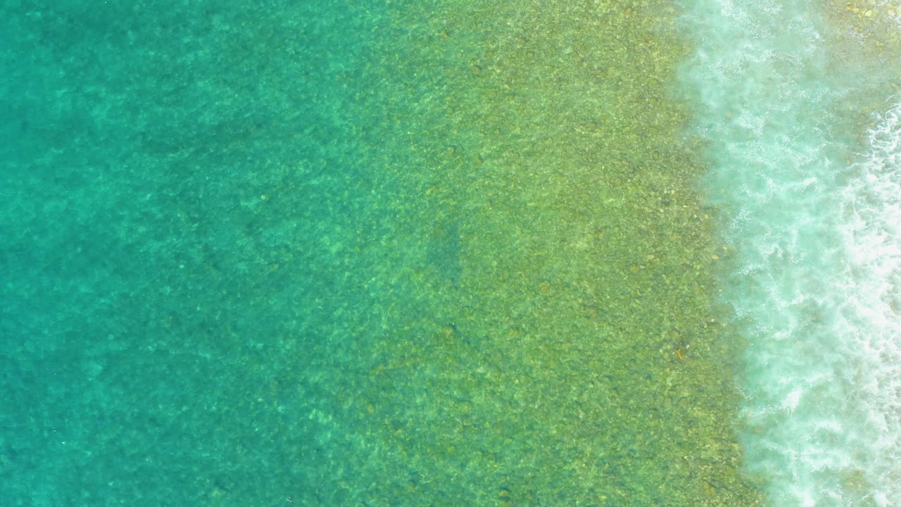 Aerial view at El Tule Beach with open turquoise water filling the left side, pale green shallows in the middle, and broken white wave foam at the right edge, no visible people.