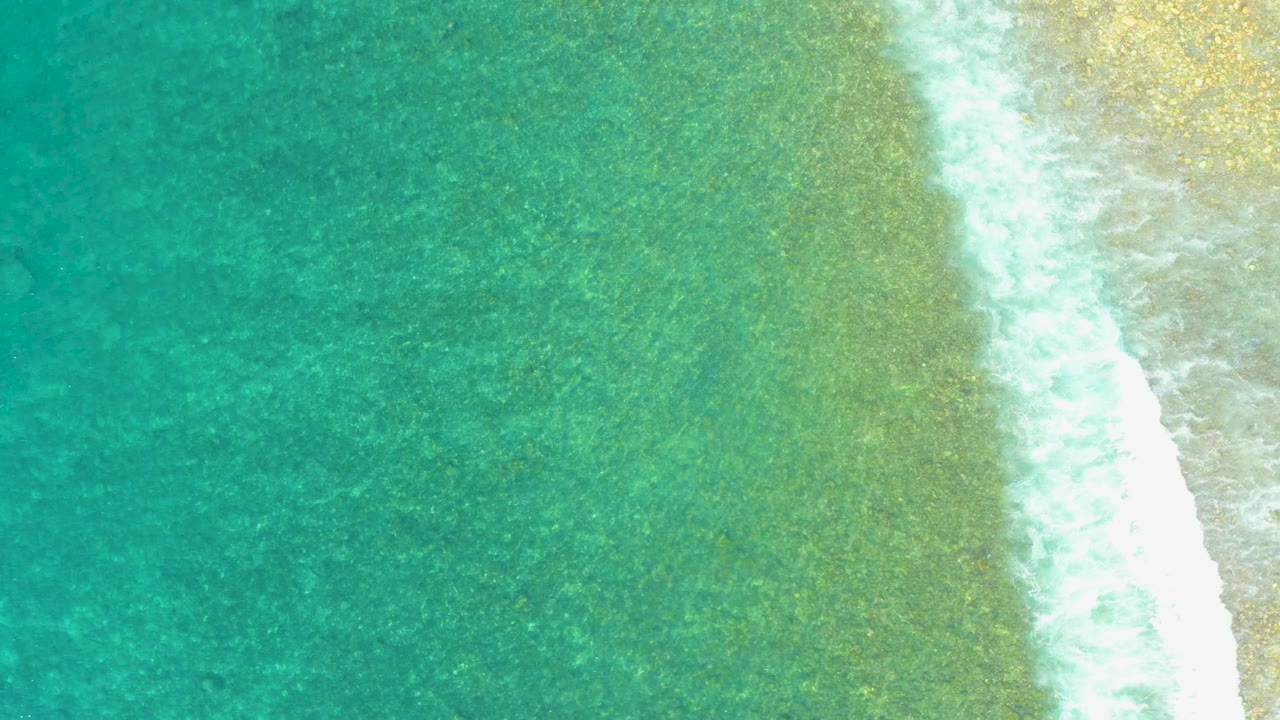Aerial frame from El Tule Beach with green-blue shallow water filling the left side, white foam breaking near the right edge, pale sand beyond it, and no visible people.