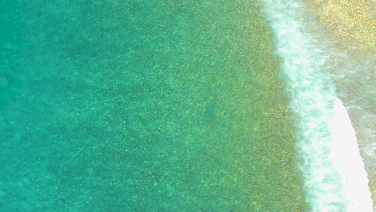 Overhead Los Cabos beach view with turquoise water, mottled green shallows, a bright curved strip of surf foam, sandy shoreline on the right, and no visible people.