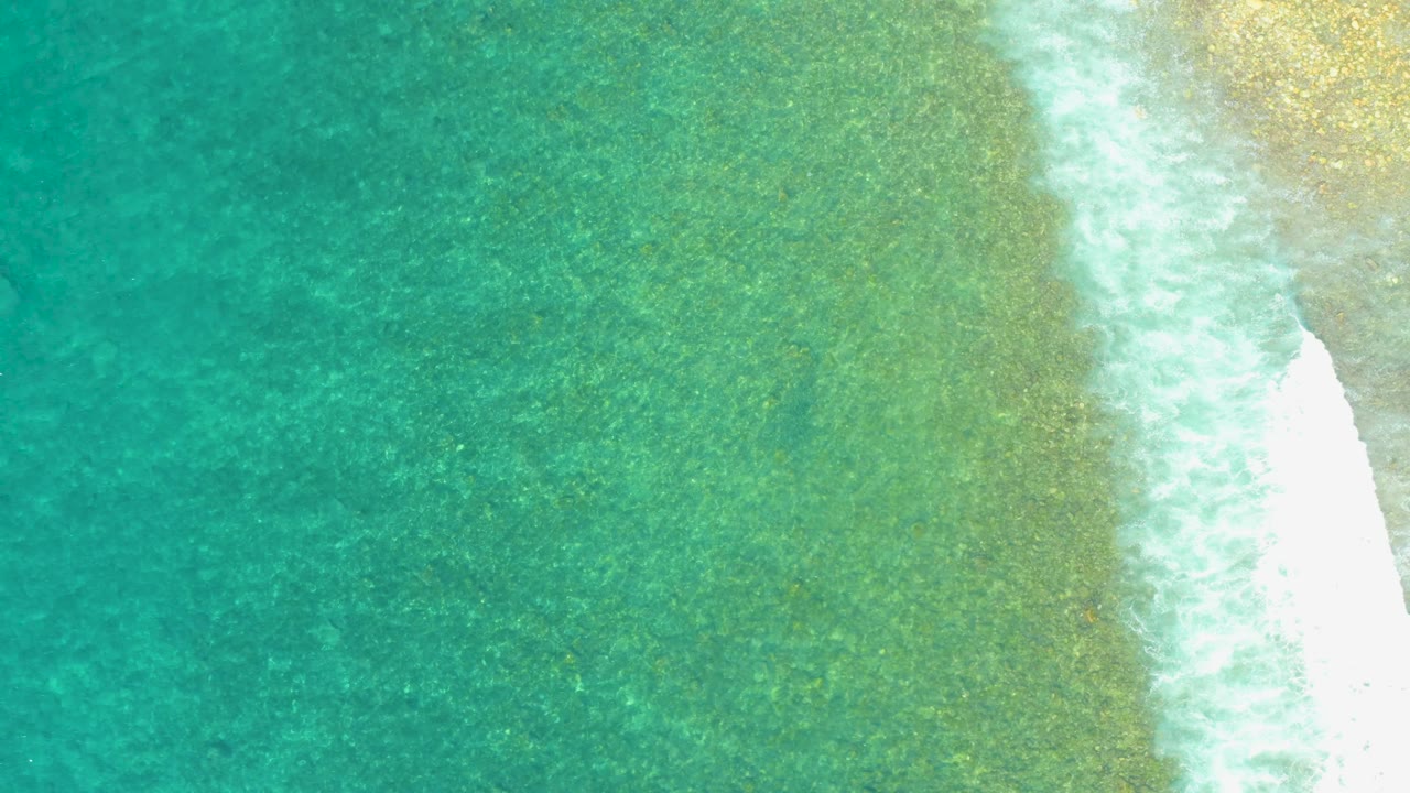 El Tule Beach aerial frame with teal water across most of the image, a diagonal white wave line near the right side, sandy beach beyond the foam, and no visible people.