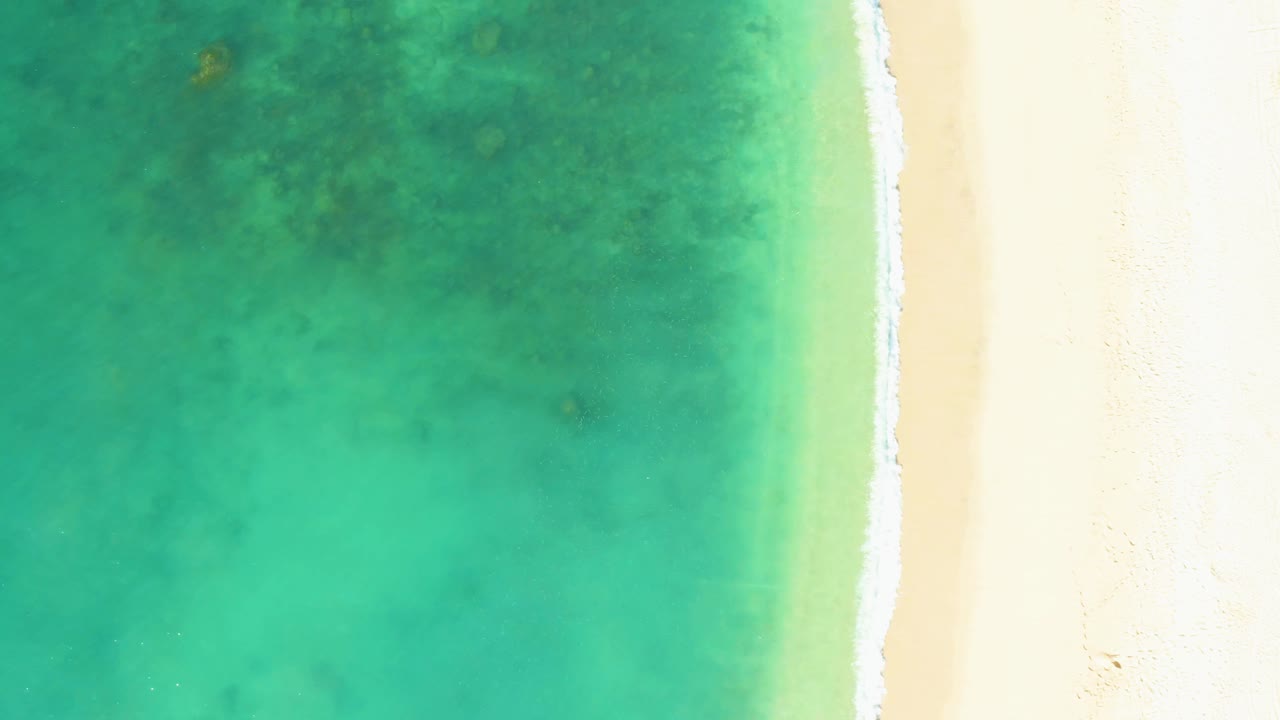 Aerial frame from El Tule Beach Los Cabos with clear green-turquoise water, a thin white surf line near the right edge, pale sand beyond it, and no visible people.