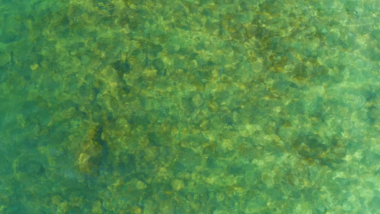 Opening overhead view of clear shallow water at El Tule Beach Los Cabos, with submerged rocks visible through green-blue ripples and no visible people.