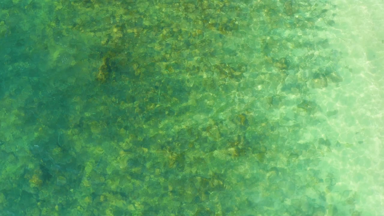 Clear turquoise-green water covers the frame at El Tule Beach Los Cabos, with ripple bands shifting across the visible stones below and no visible people.