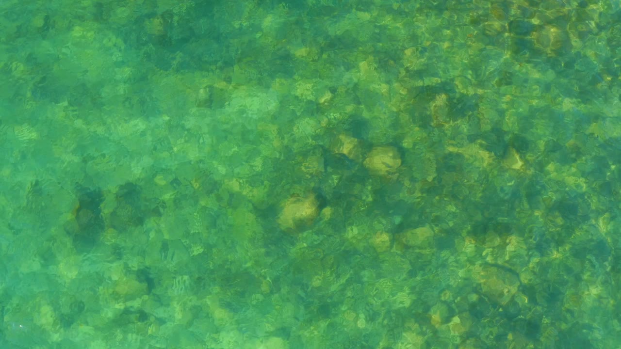 Clear shallow water at El Tule Beach in Los Cabos reveals rocks and pebbles beneath green-blue ripples, with no visible people.
