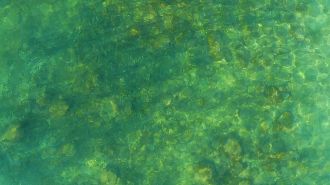 Surface ripples pass over the visible rock bed in clear shallow water at El Tule Beach in Los Cabos, with no visible people.