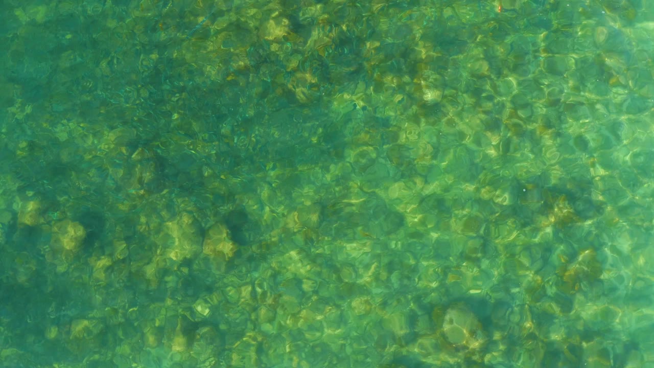 Clear shallow water at El Tule Beach in Los Cabos shows darker ripple bands crossing a green rock bed, with no visible people.