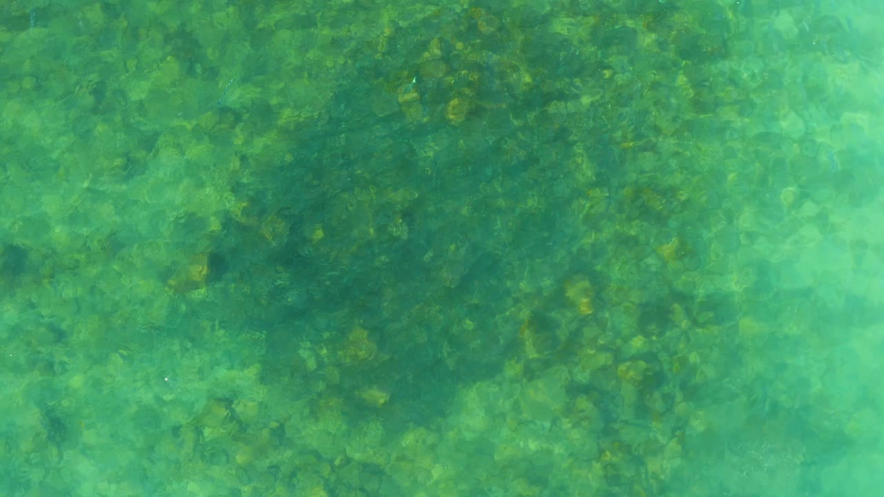 Overhead water surface at El Tule Beach Los Cabos shows mottled seabed color and shifting ripples, with no visible people.