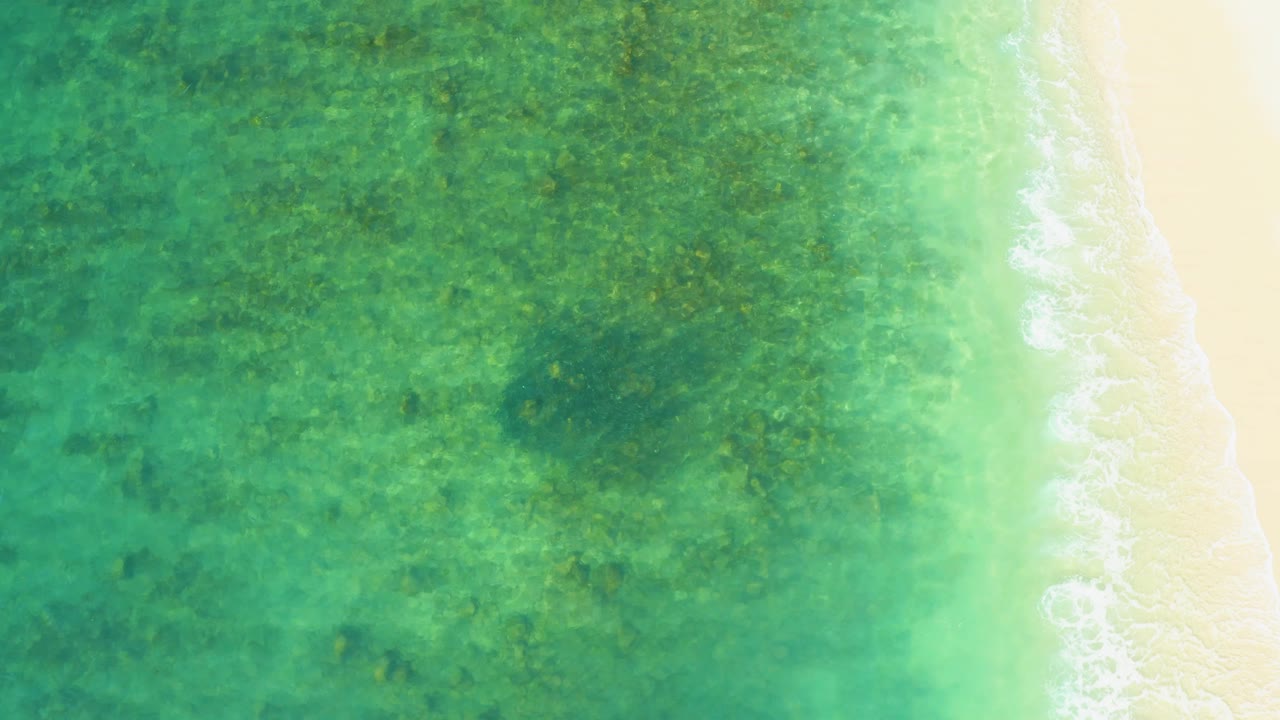Aerial frame from El Tule Beach Los Cabos with green-blue water covering a textured shallow seabed, a bright sandy strip at right, and no visible people.