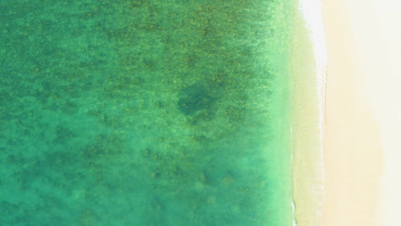 Aerial frame of clear green turquoise water beside bright sand at El Tule Beach, with the foam line close to the right edge and no visible people.