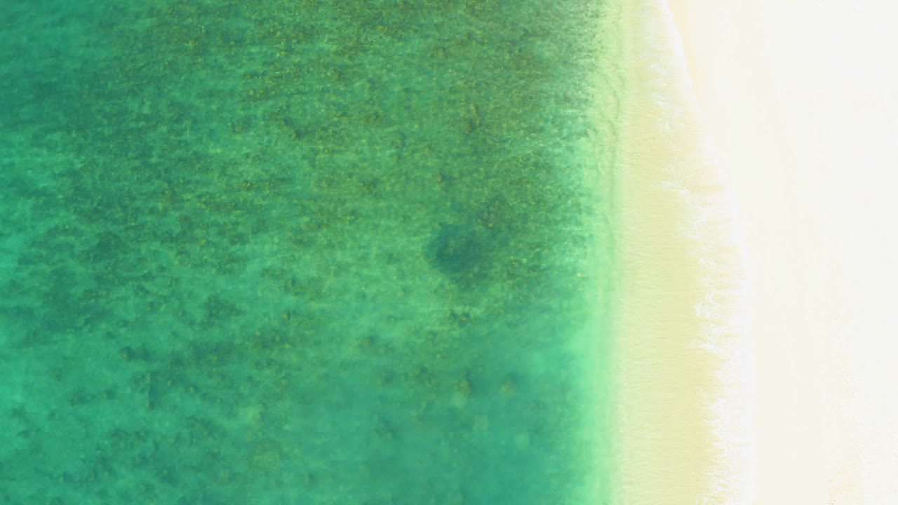 Top-down view at El Tule Beach with textured turquoise shallows, a bright sandy strip to the right, fine white wash at the waterline, and no visible people.