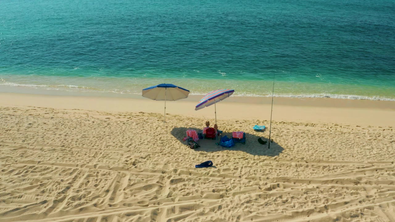 At El Tule Beach in Los Cabos, an overhead view shows two umbrellas, beach chairs, small gear, and visible people on pale sand below turquoise water.