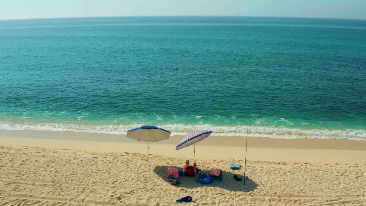 Aerial frame at El Tule Beach in Los Cabos showing two umbrellas, beach chairs, small visible people, patterned sand, and turquoise surf beyond.