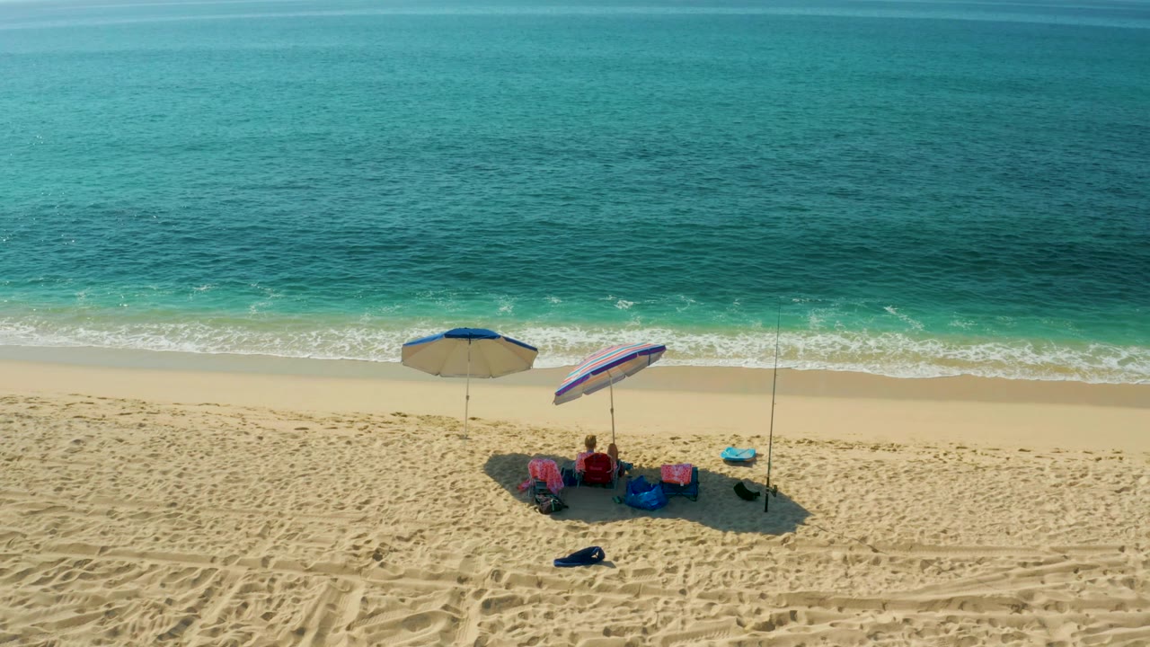 At 0:02, the aerial view shows the El Tule Beach umbrella group farther to the right, with foamy waves rolling along the upper edge of the sand.