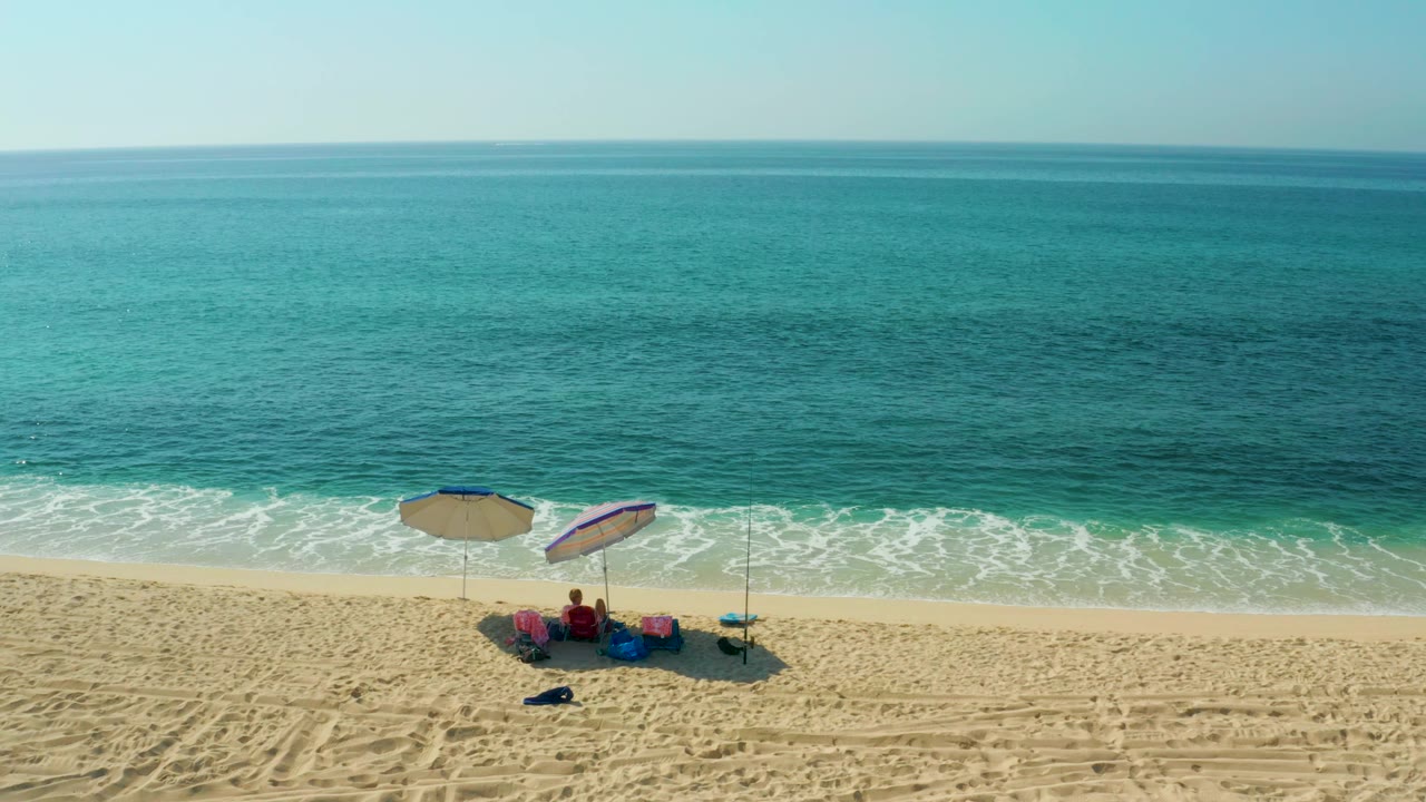 At El Tule Beach in Los Cabos, two umbrellas and seated beachgoers appear on pale sand below a wide band of turquoise ocean and a clear sky.