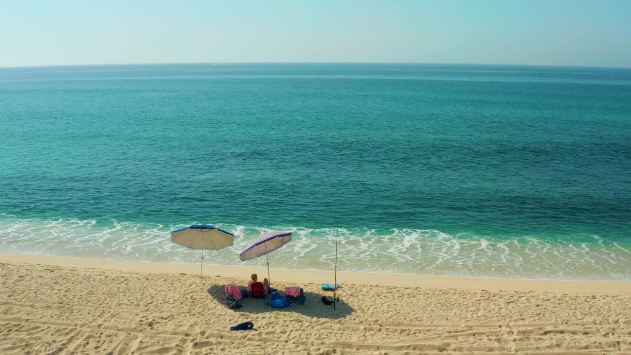 At El Tule Beach, seated people under two umbrellas sit in the lower right portion of the frame as open turquoise water stretches to the horizon.