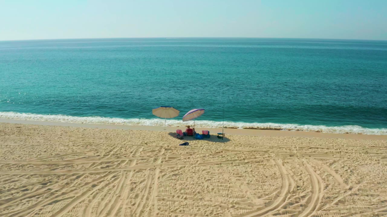 Aerial view at El Tule Beach in Los Cabos with tan sand, turquoise ocean, two umbrellas, chairs, bags, and beachgoers gathered near the waterline.