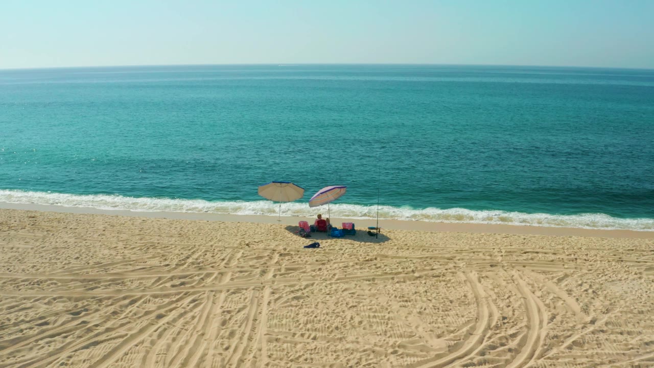 A broad aerial composition at El Tule Beach shows the turquoise sea occupying most of the upper frame, with two umbrellas, chairs, and visible beachgoers near the lower shoreline.