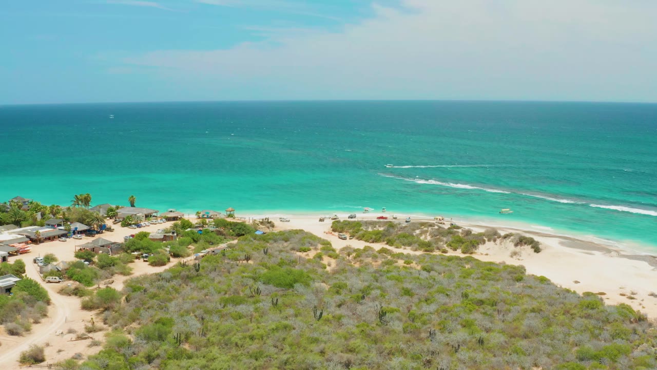 Aerial frame from Cabo Pulmo, Baja California Sur, showing turquoise ocean, white surf, pale beach, scrub-covered dunes, and no visible people.