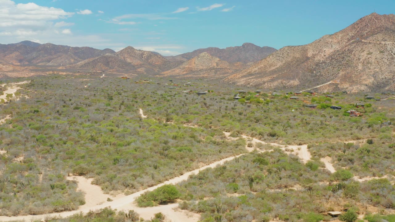 Opening aerial view over Cabo Pulmo desert terrain, with pale dirt tracks crossing low shrubs, rocky mountains on the horizon, scattered clouds, and no visible people.