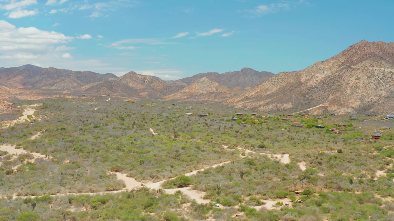 A wide aerial frame over Cabo Pulmo desert scrub shows pale dirt roads crossing the foreground, low structures in the midground, rocky mountains behind them, and no visible people.