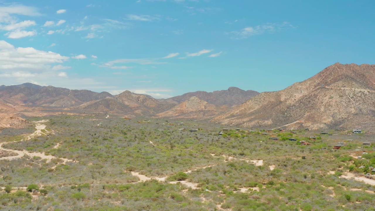 Opening frame of Cabo Pulmo, Baja California Sur, showing a wide desert valley, pale dirt roads, scattered scrub, layered mountains, blue sky, and no visible people.