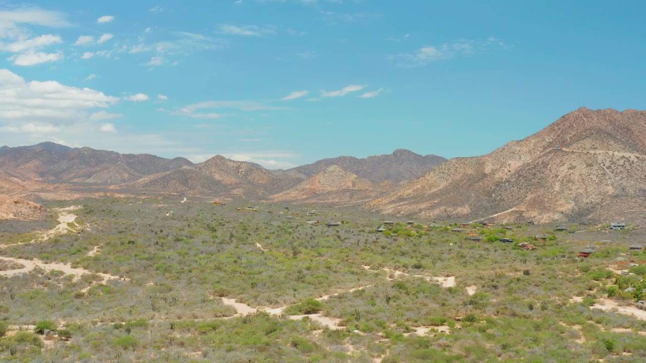 At 0:02, the Cabo Pulmo valley shows clustered green patches, pale road cuts, small distant buildings, and a larger mountain slope on the right, with no visible people.