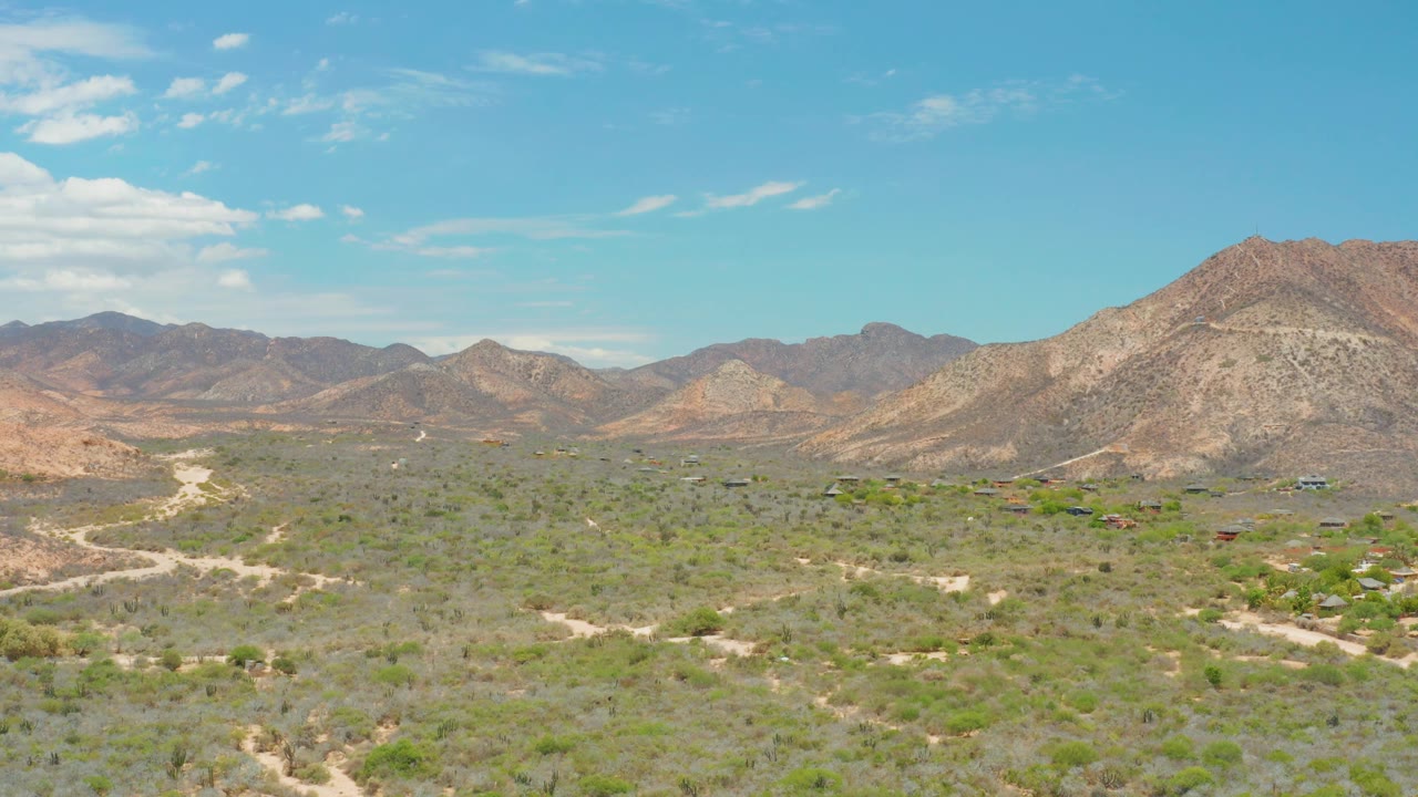 Cabo Pulmo landscape frame with dry scrubland, intersecting dirt tracks, brown mountain slopes, a few tiny structures, and no visible people.