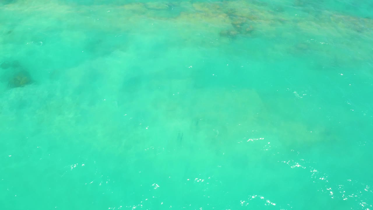 Bright turquoise water over Cabo Pulmo seabed shows subtle coral or rock patches near the top of frame, scattered white surface glints, and no visible people.