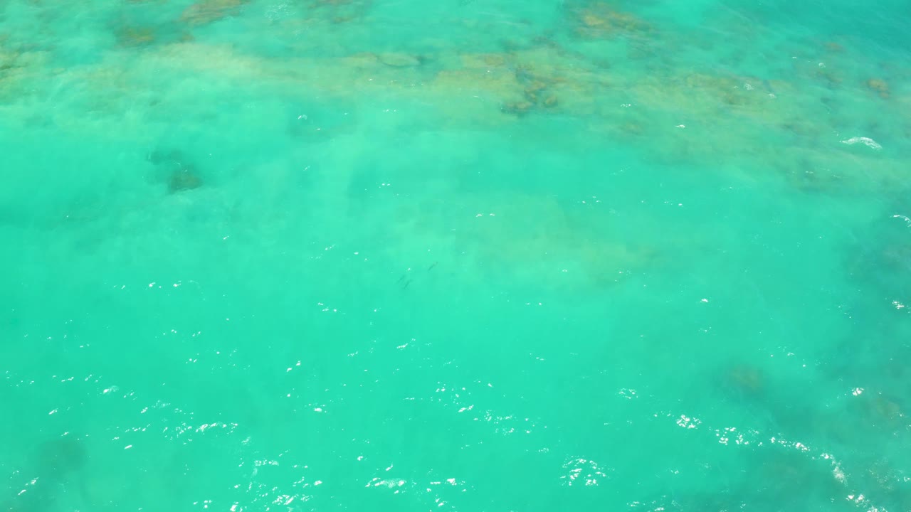 Mid-clip frame in Cabo Pulmo with a darker submerged patch near the left edge, pale seabed tones, ripples, and no visible people.