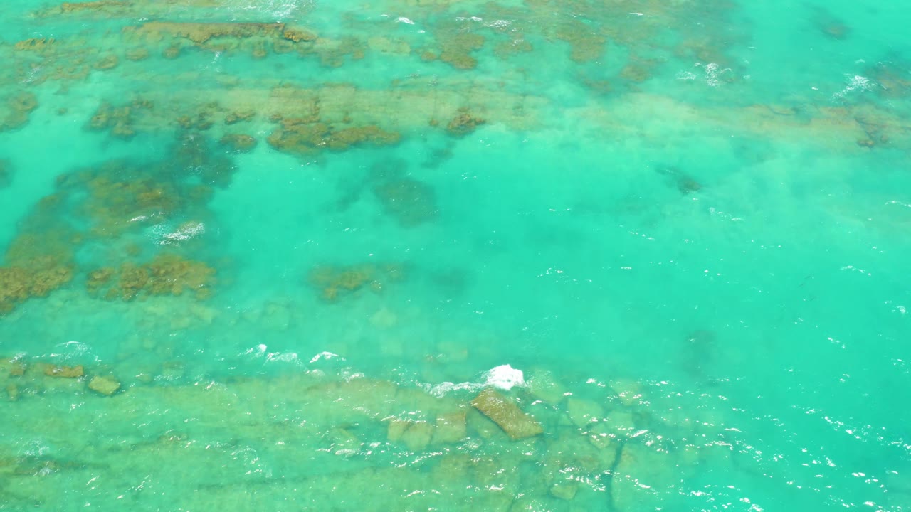 An overhead Cabo Pulmo water frame shows pale turquoise shallows, darker reef-like patches near the top, scattered rocks below, and no visible people or boats.
