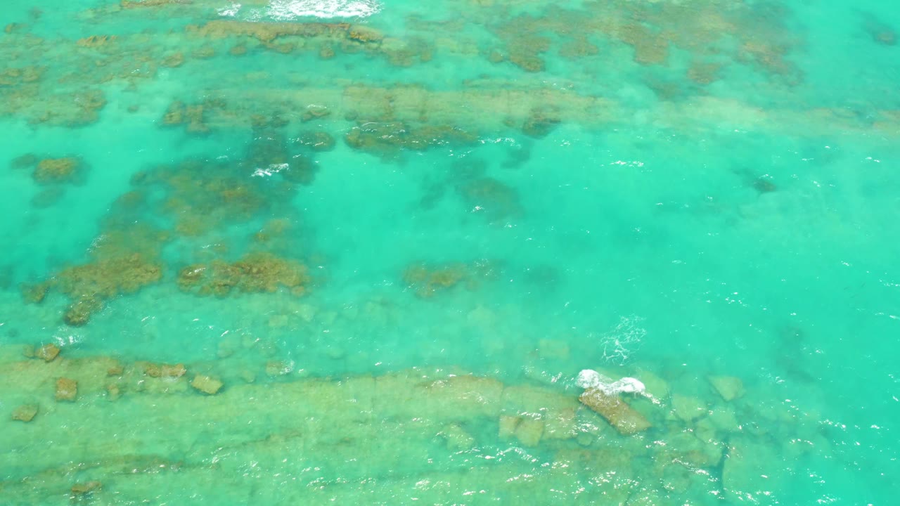 Clear turquoise water in Cabo Pulmo shows a broad submerged rock band across the lower frame, scattered darker formations above, a small foam streak, and no visible people.