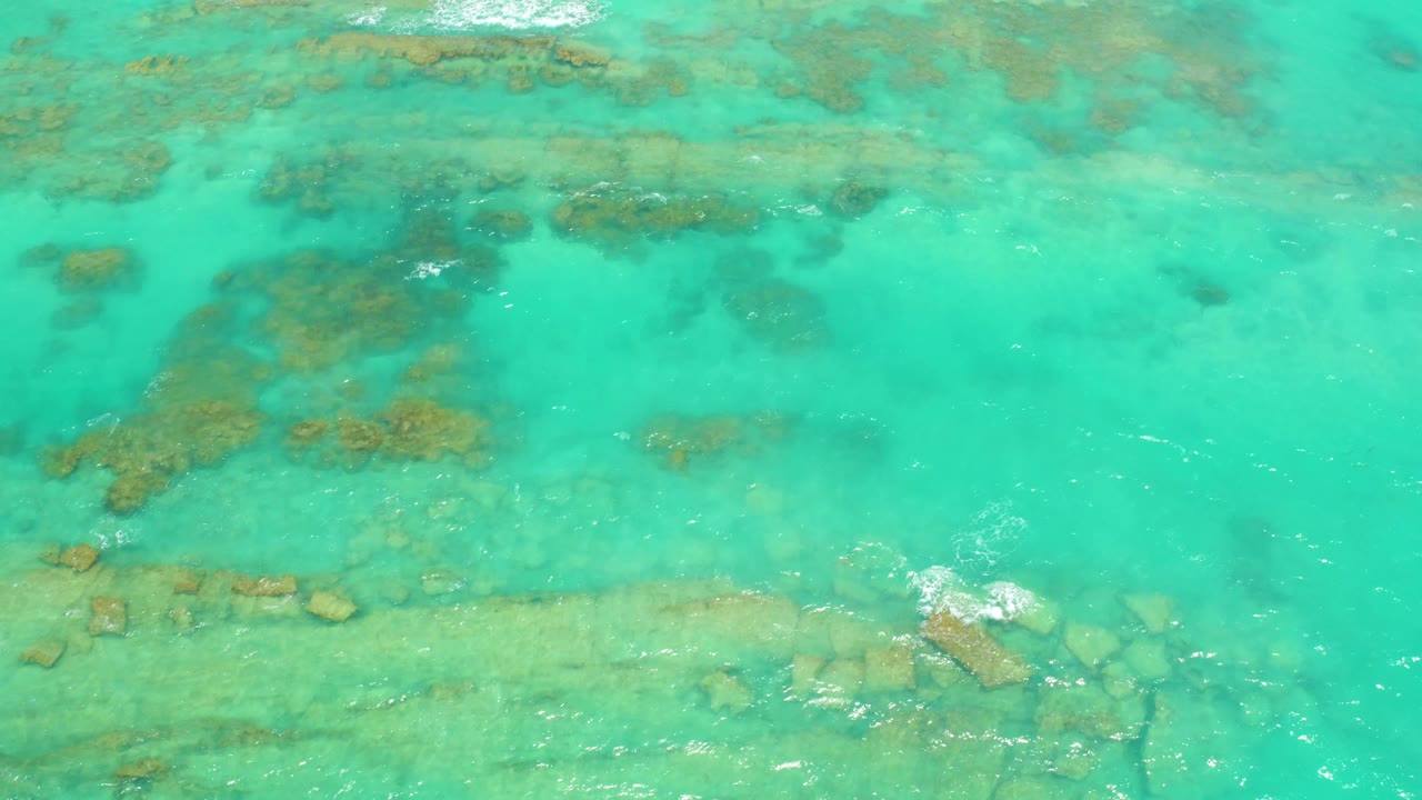 Aerial view over shallow Cabo Pulmo water with mottled underwater rocks, a pale bottom strip, white foam breaking near the right edge, and no visible people.