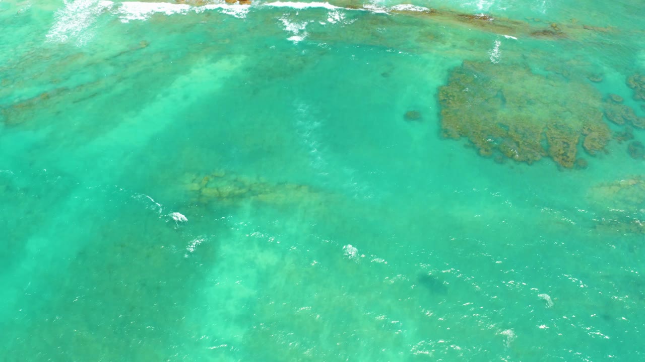 Aerial frame of clear Cabo Pulmo water with a large brown-green reef patch near the upper right and small white ripples across the surface, with no visible people.