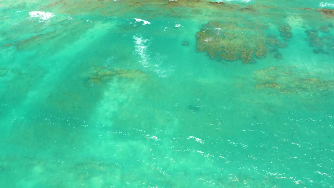 Aerial view over turquoise Cabo Pulmo ocean with a larger coral patch near the upper center, faint seabed tones below, and no visible people.