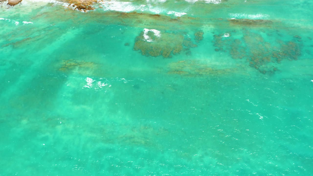 Overhead view of clear turquoise water in Cabo Pulmo, Mexico, with darker underwater rock patches, pale sand areas, white foam near the upper reef line, and no visible people.