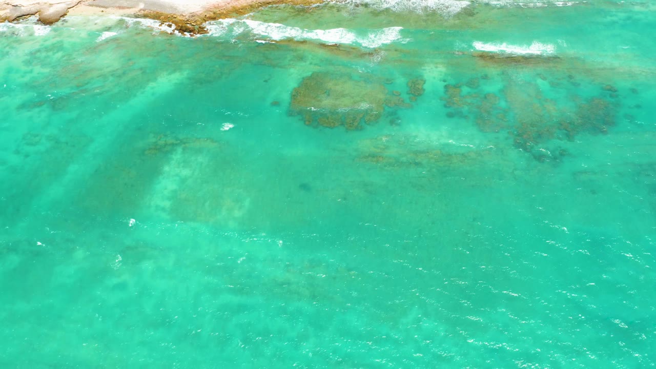 Aerial frame over Cabo Pulmo shallows with broad turquoise water, darker submerged rock or coral areas near the upper middle, breaking foam along the shore, and no visible people.