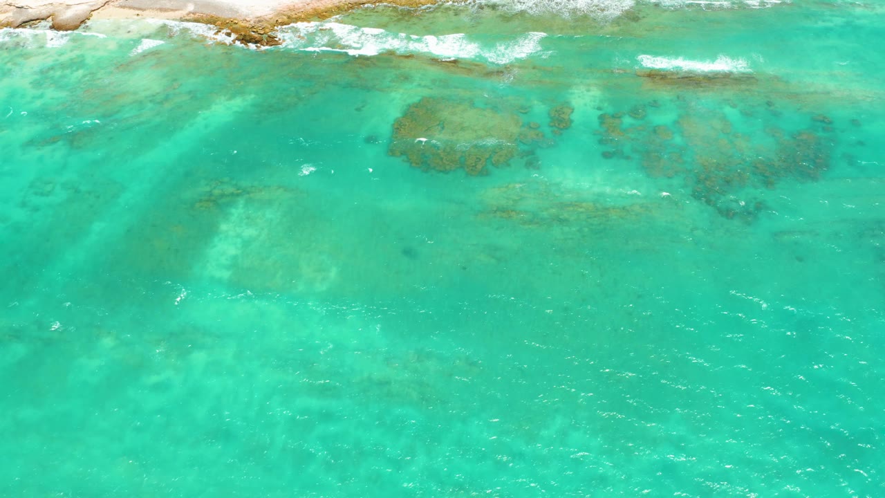 Cabo Pulmo shoreline seen from above, with a thin band of rocks at the top, white foam where waves meet the coast, green-blue water, and no visible people.