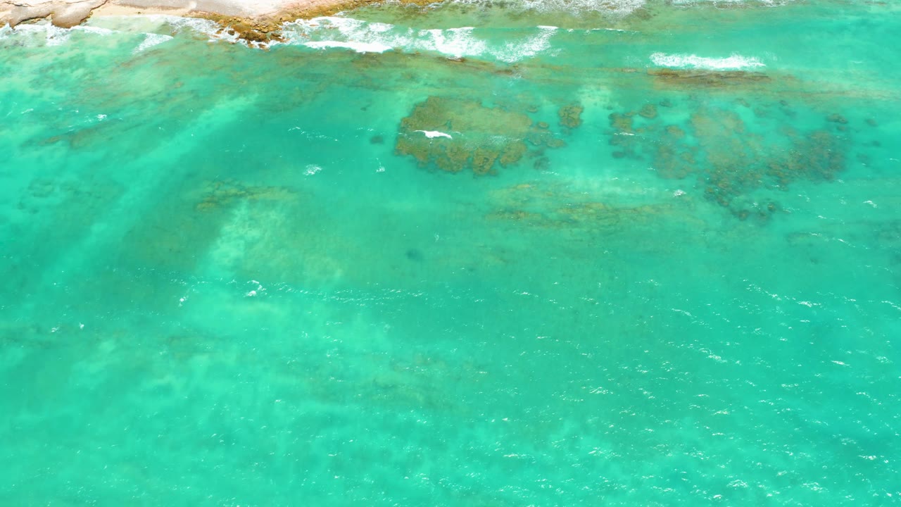 Overhead Cabo Pulmo water detail with a pale rocky shore at the top, a large darker submerged patch left of center, faint seabed patterns, white foam, and no visible people.