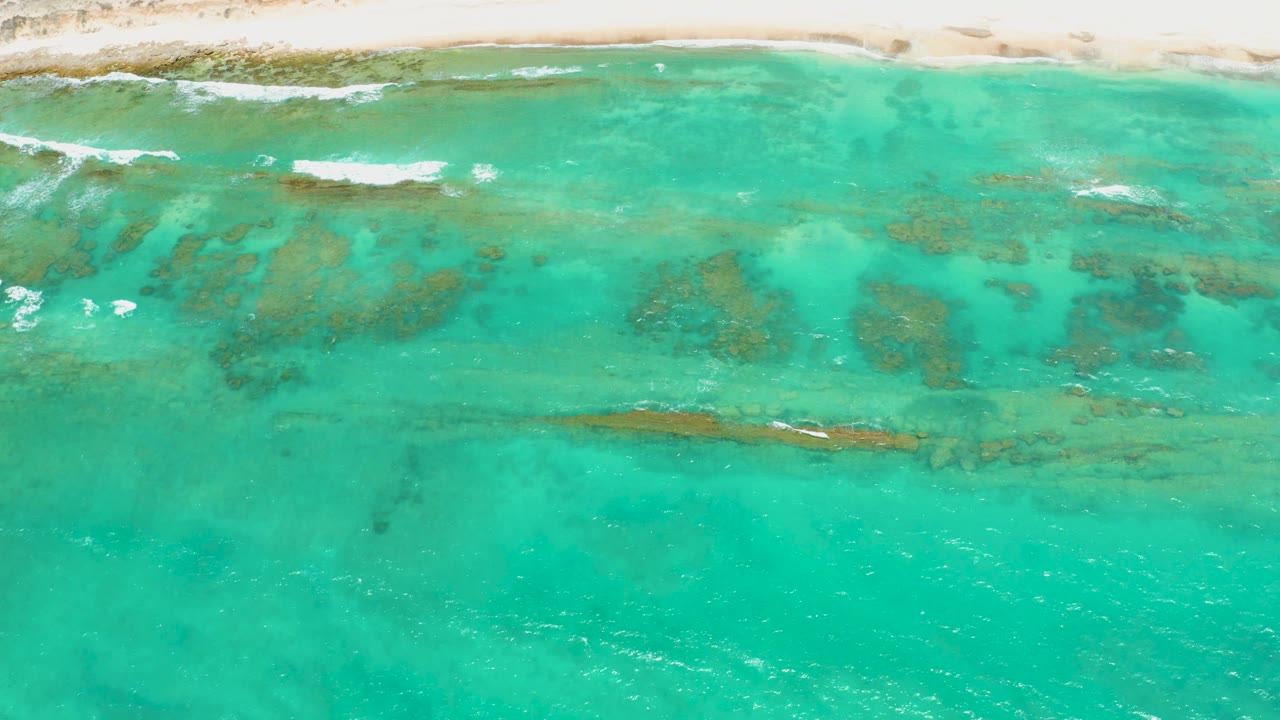 Overhead view of Cabo Pulmo shoreline with pale sand at the top, turquoise shallows below, darker seabed patches, small white wave breaks, and no visible people.