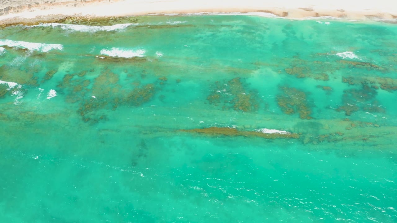 Aerial view of Cabo Pulmo shallows where white surf lines cross darker underwater patches below the sandy beach, with no visible people.