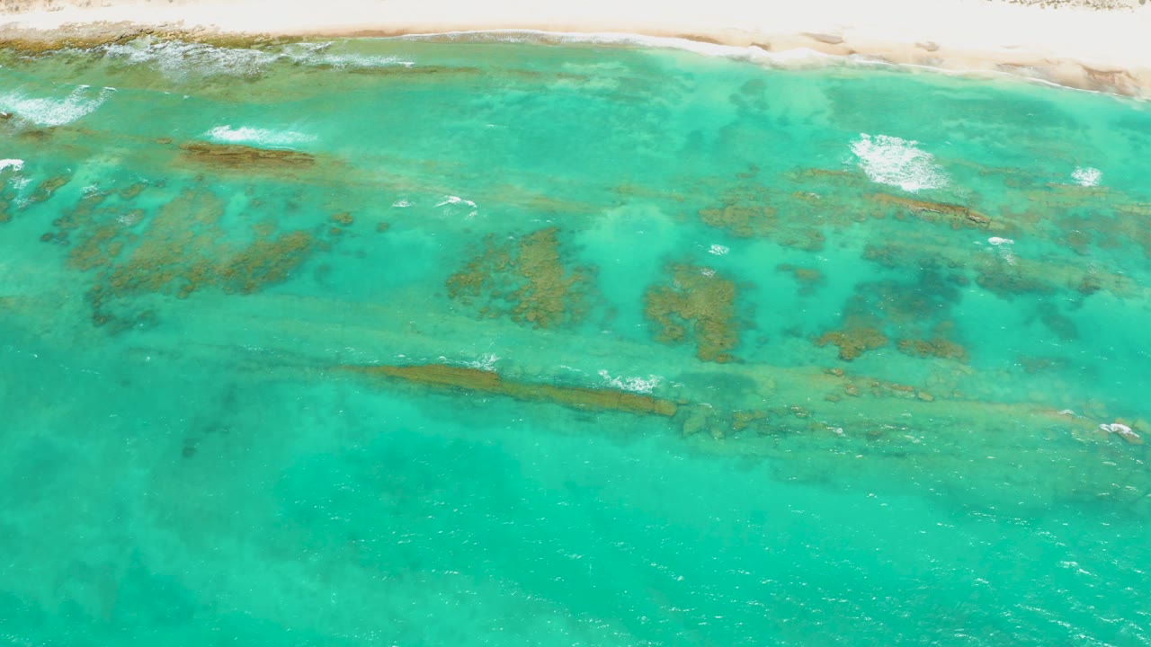 Opening overhead frame of Cabo Pulmo shallow turquoise water with darker coral reef patches below the surface, pale shoreline at the top, and no visible people.