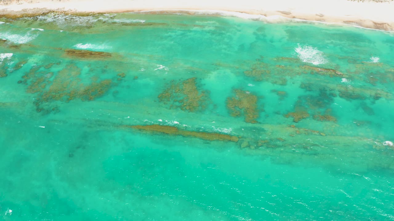 Cabo Pulmo aerial frame showing scattered coral reef patches beneath bright turquoise water, light surf near the upper shoreline, and no visible people.