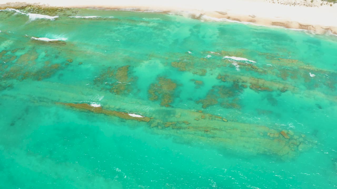 Aerial frame of clear turquoise Cabo Pulmo water with elongated submerged reef bands, pale sand along the upper edge, and no visible people.