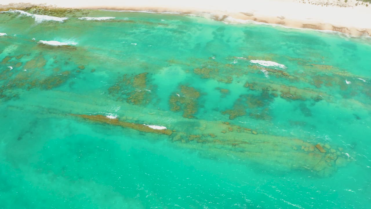 Overhead Cabo Pulmo shoreline view with turquoise shallows, darker reef patches spread across the middle, narrow white surf foam near the sand, and no visible people.