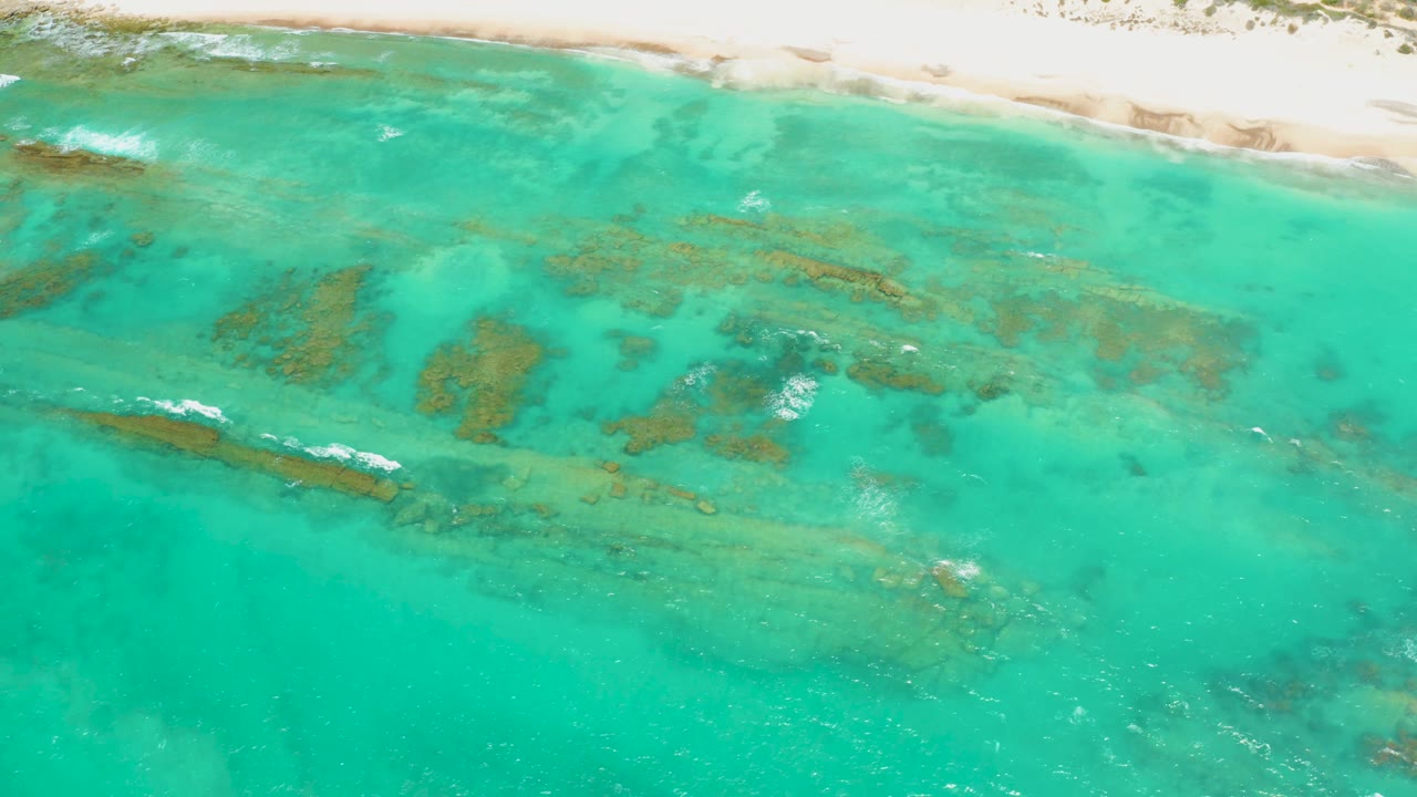 Aerial frame from Cabo Pulmo, Baja California Sur, with the sandy beach still visible along the top and brown underwater formations stretching across the turquoise water, with no visible people.